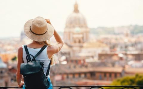 Une jeune femme, portant un chapeau de paille et un sac à dos, admire une vue panoramique sur une ville historique parsemée de dômes sous un ciel lumineux.