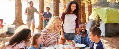 Groupe de personnes riant autour d'une table de camping avec une tente et des arbres en arrière-plan