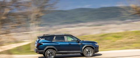 Dacia Bigster hybride bleu, vu de profil, en mouvement sur une route de campagne avec un arrière-plan flou.