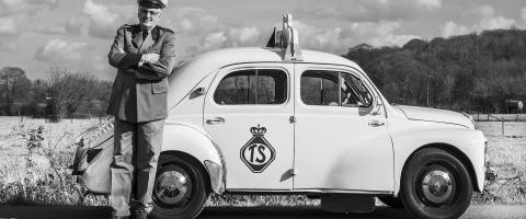 Photo noir et blanc d'un homme en uniforme avec képi appuyé contre une ancienne voiture.
