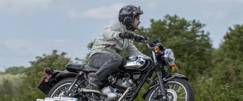 La Kawasaki Meguro S1 conduite par un motard sur fond de nature et ciel couvert.