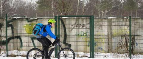 Cycliste de profil avec casque et sac à dos, roulant dans la neige, grillage vert et arbres dénudés en arrière-plan.