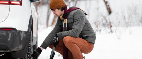 Pneus hiver : un homme accroupi vérifie la pression d'un de ses pneus, sur une voiture blanche et un paysage enneigé.