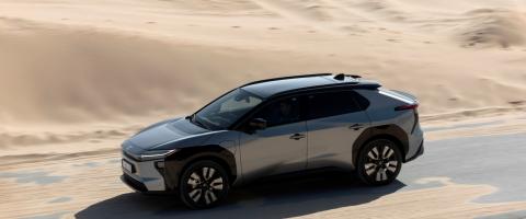 Toyota bZ4X gris vu de profil en mouvement sur une route bordée de sable.
