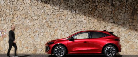 Renault Clio rouge de profil, stationnée devant un mur en pierre, avec un passant flou au premier plan.