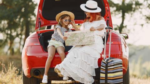 Mère et fille préparent leur voyage sur la route, assises à l’arrière d’une voiture rouge avec une carte et une valise à leurs côtés.