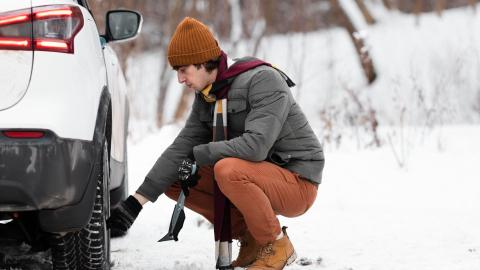 Pneus hiver : un homme accroupi vérifie la pression d'un de ses pneus, sur une voiture blanche et un paysage enneigé.