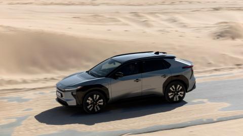 Toyota bZ4X gris vu de profil en mouvement sur une route bordée de sable.