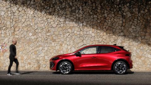 Renault Clio rouge de profil, stationnée devant un mur en pierre, avec un passant flou au premier plan.