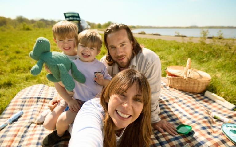 Une famille souriante prend un selfie lors d'un joyeux pique-nique ensoleillé au bord d'un lac, profitant de la nature luxuriante.