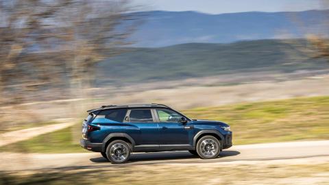 Dacia Bigster hybride bleu, vu de profil, en mouvement sur une route de campagne avec un arrière-plan flou.